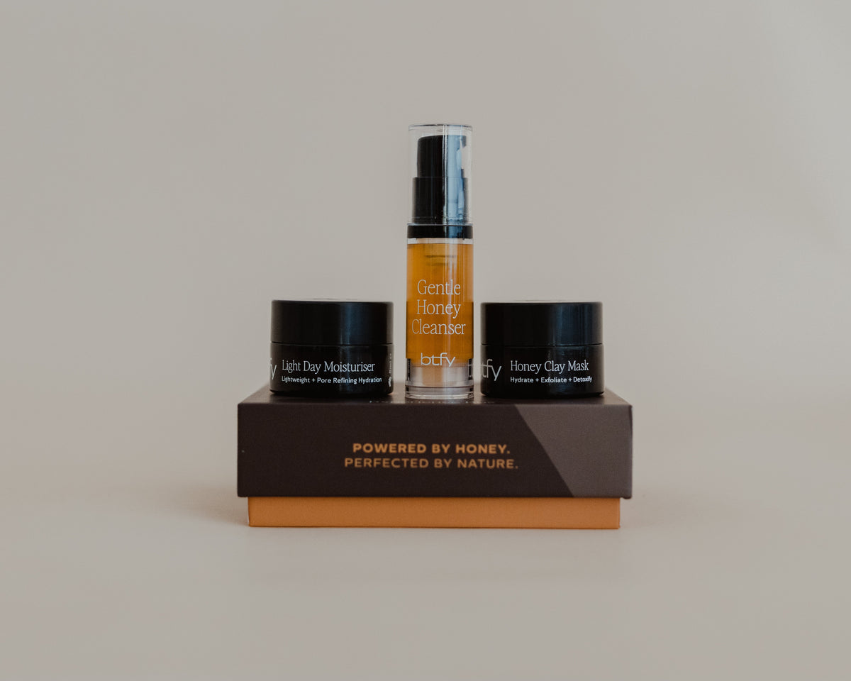 Three skincare products from Beauty by Honey are displayed on a dual-tone brown and orange stand against a neutral beige background. The products include a Honey Glow Cleanser spray bottle, a Light Hydrating Moisturizer jar, and a Honey Clip Mask jar.