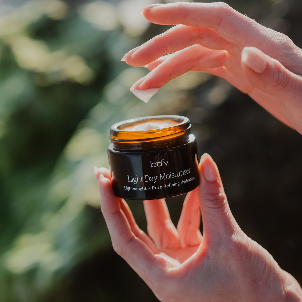 A person's hands holding an amber glass jar of btfy Light Day Moisturiser, with a cotton swab being lifted from the open jar, set against a blurred outdoor background.