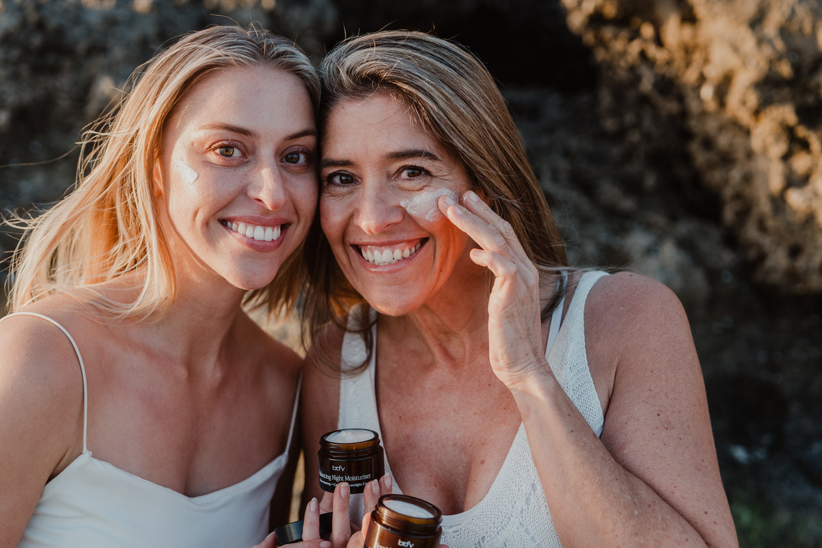 Two women smiling outdoors in front of a rocky background, with one applying BTFY moisturiser to her face and both holding amber jars. The scene is warm, golden-hour lit and clearly lifestyle-oriented.