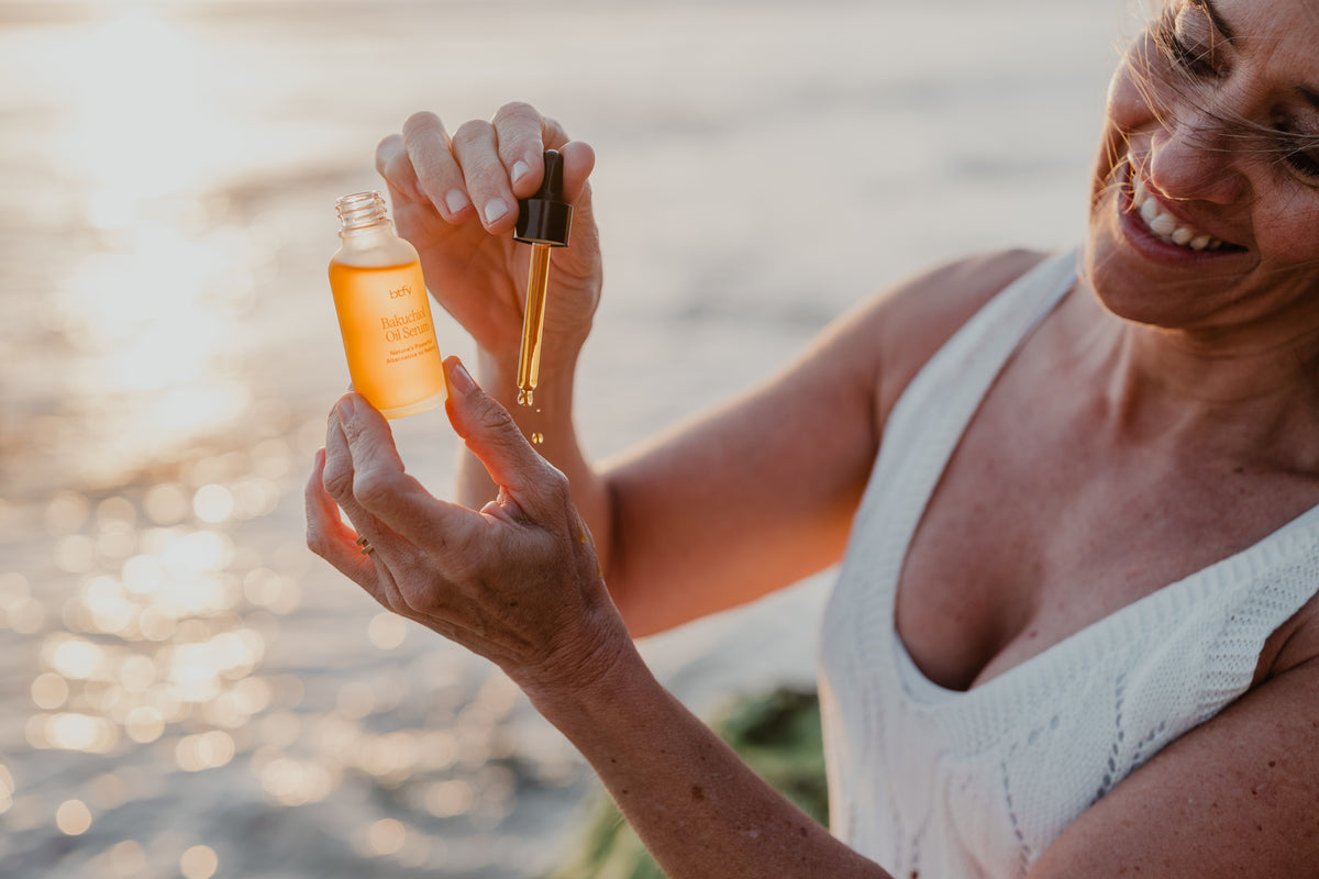 A woman outdoors by the water holds a BTFY Bakuchiol Oil Serum amber dropper bottle and a gold dropper, with warm sunlight reflecting on the water in the background.