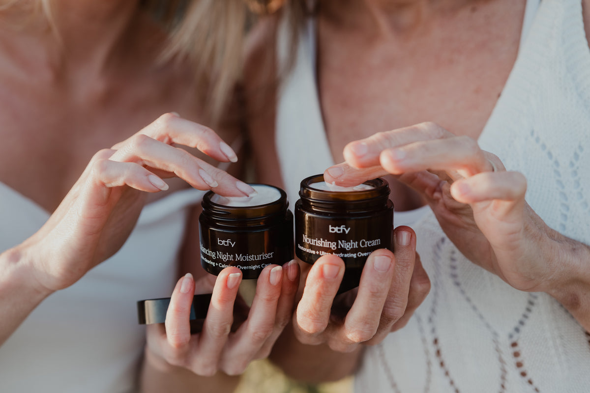 Two people holding two dark amber jars of BTFY skincare, one labeled 'Balancing Night Moisturiser' and the other 'Nourishing Night Cream', with white cream visible on the lids.