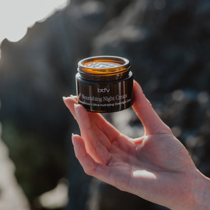 A hand holding a small amber glass jar of btfv Nourishing Night Cream against a blurred outdoor rocky background.