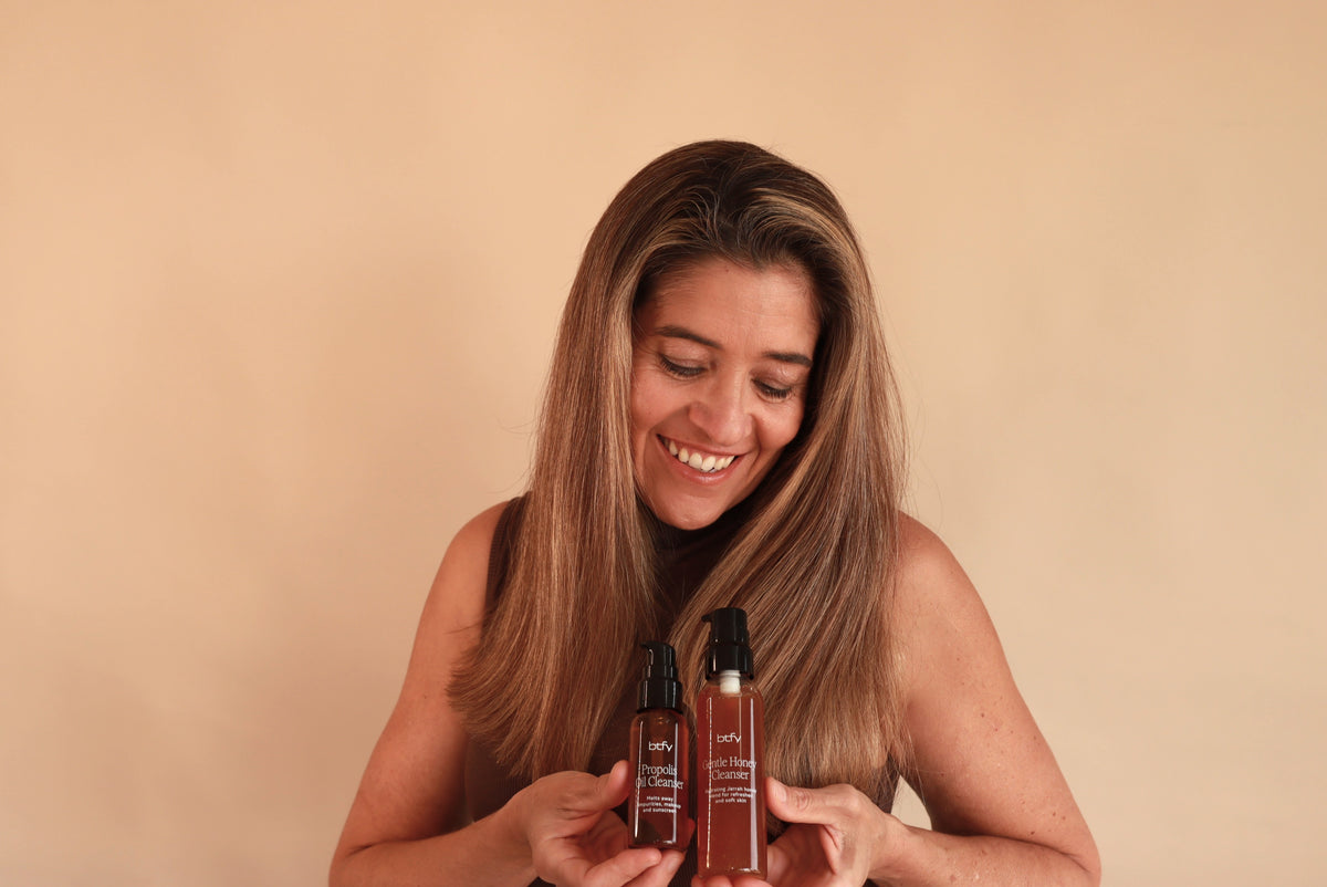A smiling woman with long light brown hair holds a BTFY Propolis Oil Cleanser and Gentle Honey Cleanser against a solid beige background.