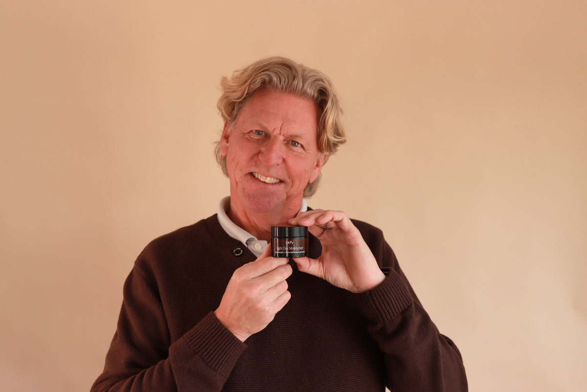 A smiling man with blond hair wearing a brown sweater holds up a small dark jar or container against a plain beige background.