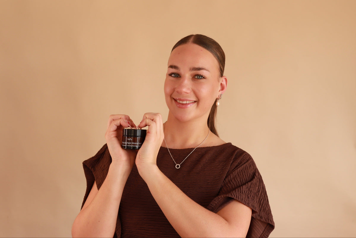 A woman with her hair pulled back, wearing a dark brown textured top and a delicate necklace, holds up a small BTFY Balancing Night Moisturiser against a plain beige background.