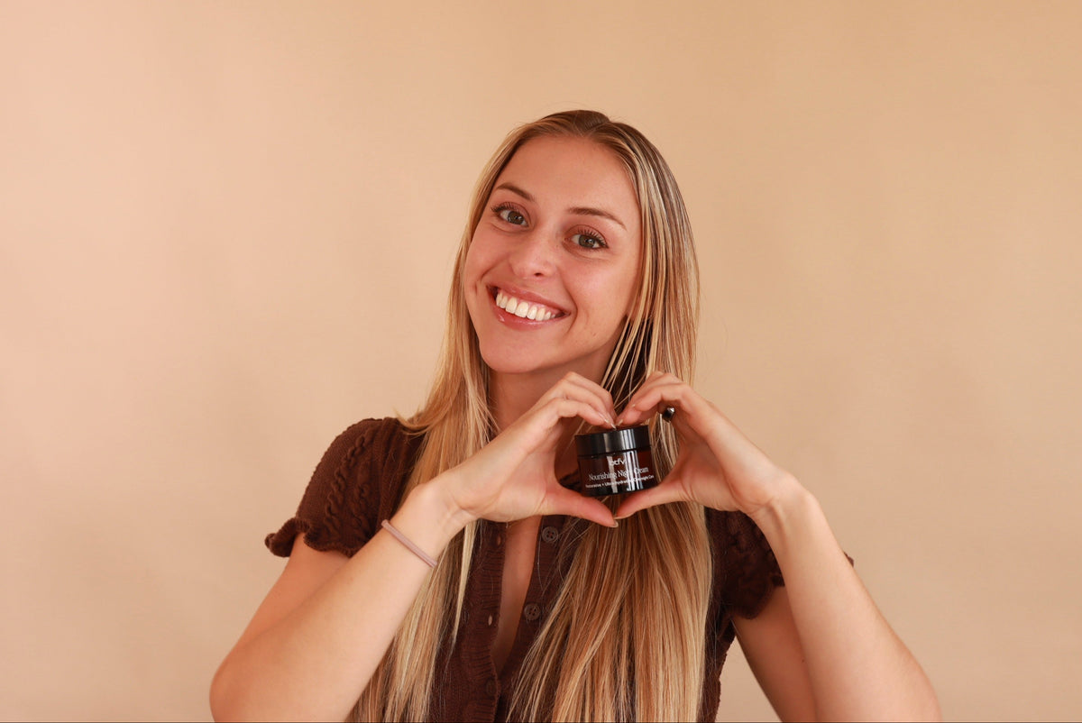 A smiling woman with long blonde hair holds a BTFY Nourishing Night Cream between her hands, forming a heart shape with her fingers against a plain beige background.