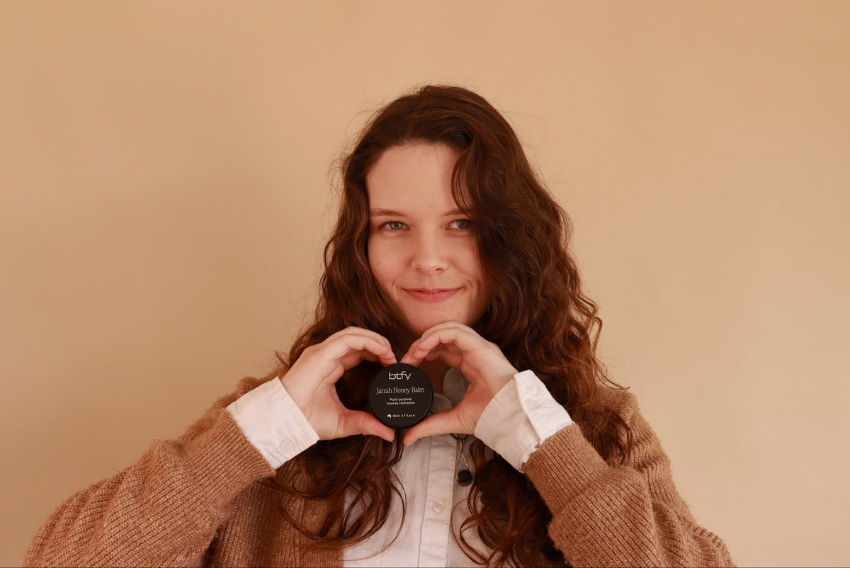 A woman with long wavy brown hair holds the BTFY Jarrah Honey Balm in front of her chest, forming a heart shape with her hands against a solid beige background.